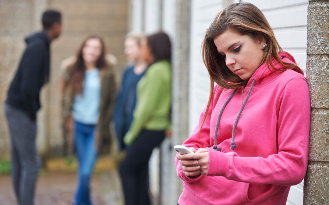 teinityttö selaa puhelinta yksin, taustalla nuorten ryhmä juttelee keskenään.