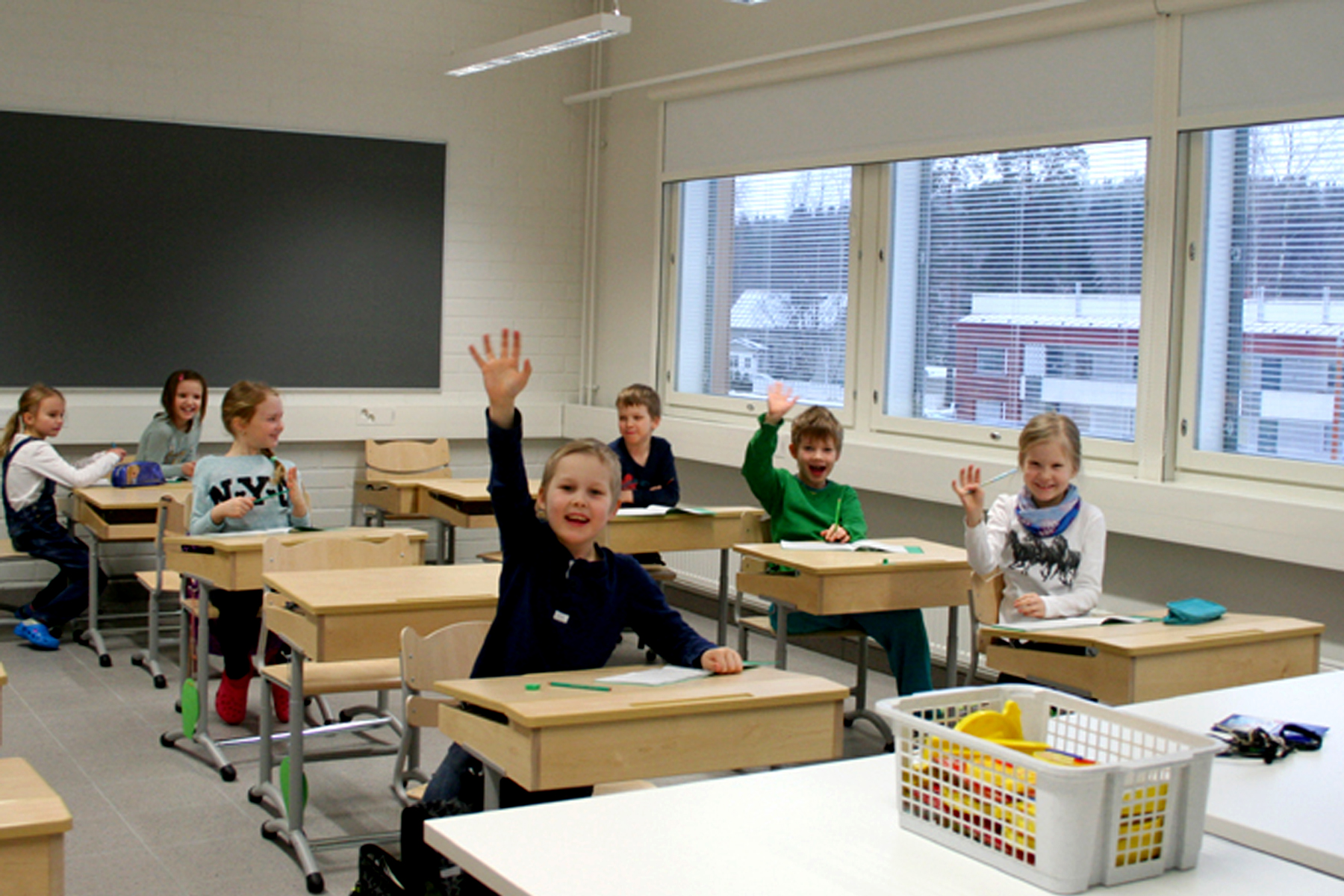Kinkomaan koulun luokkahuoneessa oppilaat istuvat pulpettiensa ääressä ja kolme oppilasta viittaa ja hymyilee.
