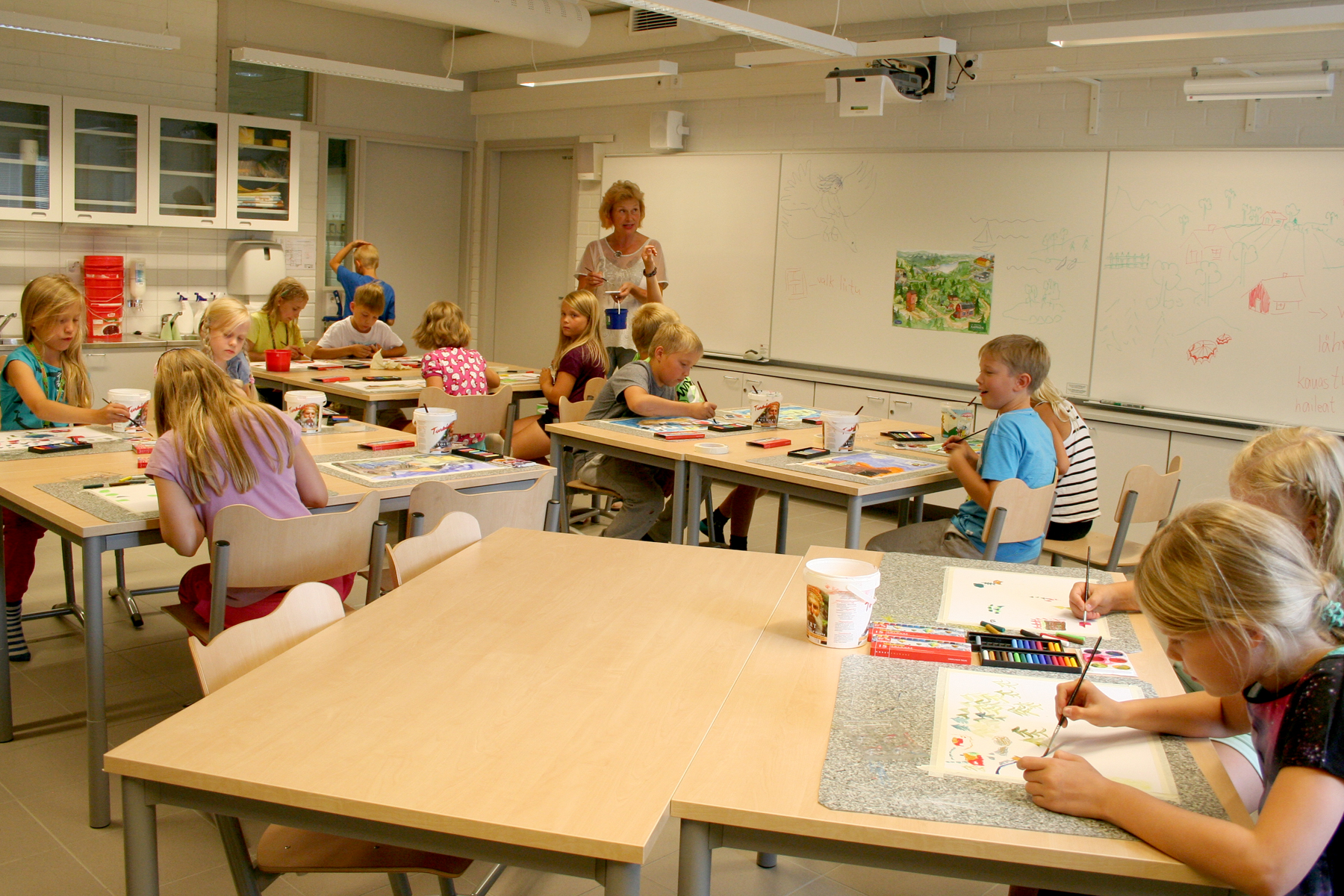 Mäkelänmäen koulun luokassa oppilaat maalaavat vesiväreillä pulpettiensa ääressä, ja opettaja seisoo luokan edessä.